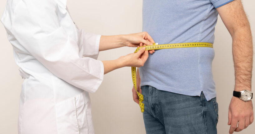 Nutritionist measuring overweight man's waist with tape in clinic, closeup